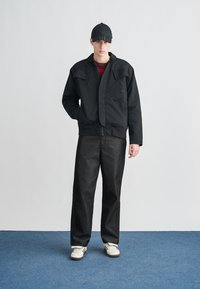 Černá bomber bunda s nášivkami na kapsách, nošená přes tmavě vínové pruhované tričko a doplněná volnými tmavými džíny a bílými teniskami. Černá čepice.