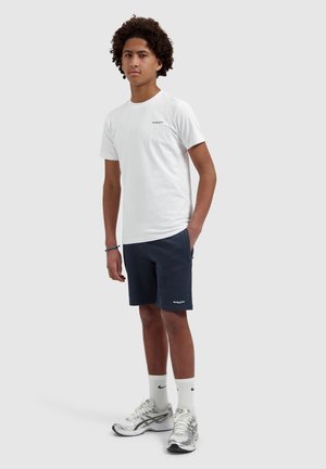 Teenage boy with curly hair wearing a white t-shirt, navy shorts, white Nike socks, and silver running shoes, standing with one hand in pocket.