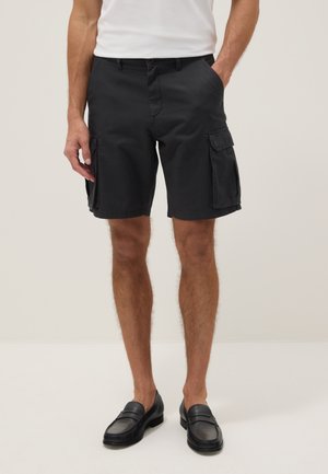 Uomo che indossa pantaloncini cargo neri con tasche laterali, camicia bianca e mocassini neri in pelle, in piedi su uno sfondo chiaro e uniforme.