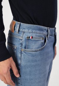 Vaqueros de mezclilla azul claro con un parche de cuero, cinco bolsillos y un pequeño acento de rayas rojas, blancas y azules en el bolsillo delantero.