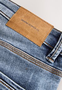 Zbliżenie na niebieskie dżinsy z pętlą na pasek, przeszyciami i brązową skórzaną metką z nadrukiem "DENIM SUPPLY CO.".