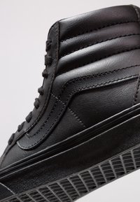 Czarne trampki typu high-top z wyś cushioninginającym kołnierzem, teksturowanym bocznym paskiem, czarnymi sznurówkami i podeszwą w wzór waflowy, na neutralnym tle.