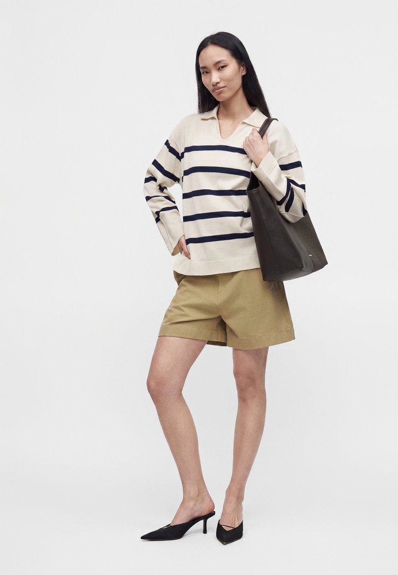 Femme debout, une main dans la poche, portant un short beige, un pull crème à rayures noires, des talons noirs, et tenant un grand sac cabas noir.