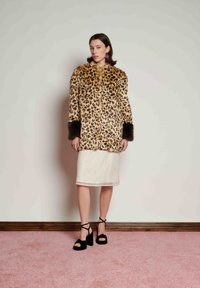 Mulher vestindo casaco de pele com estampa de leopardo e punhos escuros, saia branca até ao joelho, brincos roxos e saltos plataforma pretos, em pé sobre um tapete rosa.