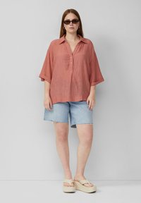 Koraalkleurig, textuurshirt met een knoopsluiting aan de voorkant en opgerolde mouwen, gecombineerd met lichtblauwe denimshorts en beige platformsandalen.