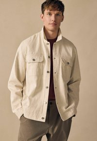 Jachetă ușoară bej cu două buzunare pe piept, cu butoane tip snap și o croială lejeră, asortată cu o cămașă bordeaux și pantaloni gri texturat.
