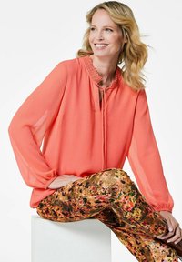 Korallfarbige Bluse mit langen, luftigen Ärmeln und einem Rüschenkragen. Kombiniert mit gemusterten, strukturierten Leggings in Erdtönen und lebhaften Blumenmustern.