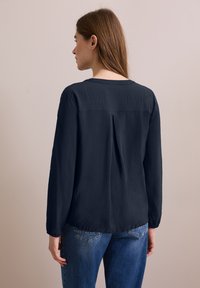 Navy blauwe blouse met lange mouwen in lichtgewicht stof, voorzien van een rugplooien, ronde halslijn en licht gatherende manchet. G gedragen met blauwe jeans.