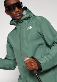 Vandeniui atspari žalia striukė su kapišonu iš The North Face; turi priekinius užtrauktukus, reguliuojamus raištelius ir subtilią logotipą ant krūtinės ir rankovės.