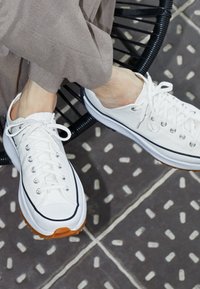 Vita canvas sneakers med svarta detaljer och silverfärgade öglor, med en texturerad orange gummisula. Bärs med ljusgrå byxor.
