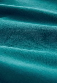 Tyrkysová látka se hladkým povrchem, s jemnými diagonálními žebry. Materiál vypadá měkce a poddajně, krásně splývá v jemných vlnách.