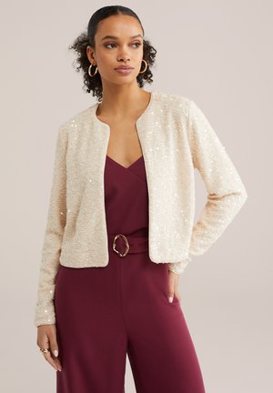 Vrouw met krullend haar die gouden oorringen draagt, een crème zijden jasje met pailletten en een mouwloze bordeauxrode jumpsuit met een bijpassende riem.