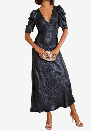 Žena v lesklých černých midi šatech s nafouknutými rukávy, držící kovové psaníčko a obuté do sandálů na vysokém podpatku se zavazováním.