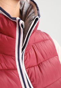 Gros plan sur une personne portant une veste matelassée rouge avec un col zippé ornée de bordures rayées blanches et bleu marine.