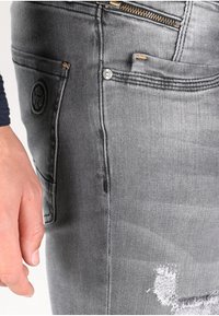 Jeans de mezclilla gris con un acabado desgastado, que cuentan con un bolsillo con cremallera y un parche redondo con logo. Diseño clásico de cinco bolsillos.