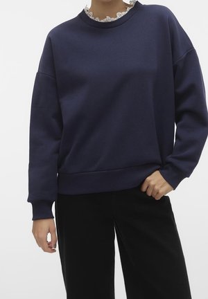 Femme portant un sweat-shirt à manches longues bleu marine par-dessus un haut avec un col en dentelle blanche, assorti à un pantalon noir, debout avec une main dans la poche.