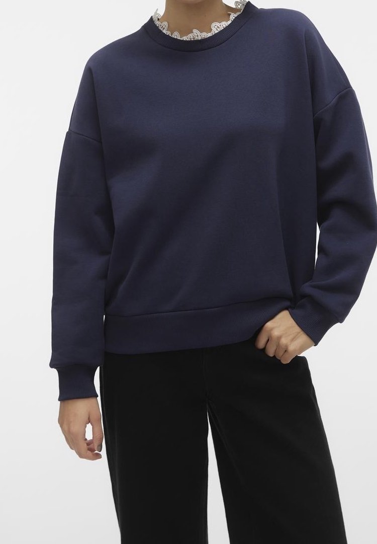 Femme portant un sweat-shirt à manches longues bleu marine par-dessus un haut avec un col en dentelle blanche, assorti à un pantalon noir, debout avec une main dans la poche.