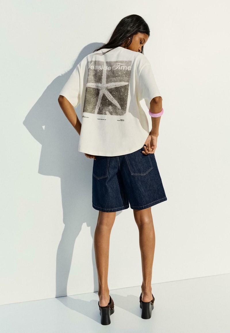 Femme portant un t-shirt blanc oversize avec un imprimé étoile de mer et un texte, un short en jean foncé, des sandales noires à talons et un bracelet rose, face au mur.