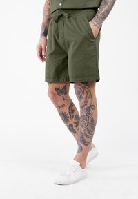 Uomo con tatuaggi floreali e di farfalla, che indossa pantaloni corti verdi oliva e sneakers bianche, in piedi davanti a uno sfondo bianco.