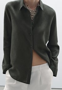 Chemise à manches longues et boutons de couleur vert foncé en tissu léger, présentant une coupe décontractée et un ourlet arrondi, portée avec un pantalon blanc.