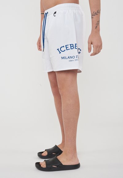 Shorts de bain blancs avec le texte bleu "ICEBEG MILANO IT". Dotés d'une taille élastique avec cordon de serrage et de poches latérales. Portés avec des sandales noires.