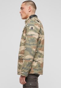 Geacă de camuflaj din bumbac cu un accent negru pe umăr. Are un buzunar frontal și detalii cu nasturi, prezentată pe un model din profil.