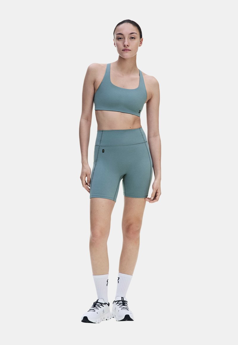 Femme debout portant un soutien-gorge de sport bleu sarcelle et un short cycliste taille haute, des chaussettes blanches et des chaussures de course blanches sur un fond clair uni.
