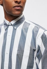 Ριγέ πουκάμισο με εναλλασσόμενες λεπτές κάθετες ρίγες σε γκρι και λευκό, γιακά με κουμπιά και τσέπη στο μπροστινό μέρος, κατασκευασμένο από απαλό ύφασμα.