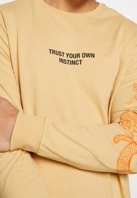 Beige Langarmhemd mit dem Aufdruck "VERTRAUE DEINEN EIGENEN INSTINKTEN" in Schwarz auf der Vorderseite und orangefarbenem Schlangen-Stickerei am Ärmel. Weiche Textur.