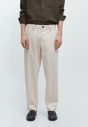 Uomo che indossa pantaloni beige a gamba dritta, mocassini neri e una camicia marrone scuro a maniche lunghe con le maniche arrotolate fino agli avambracci.