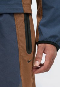 Nike μπουφάν με μπλε και καφέ χρώματα, στην επιφάνεια με λεία υφή, αντίθετη ραφή, μαύρες λεπτομέρειες και με φερμουάρ στο πλάι.