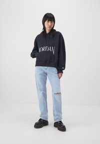 Sudadera negra con el logo "JORDAN", bolsillo tipo canguro, combinada con jeans desgastados de color azul claro y zapatos negros de cordones gruesos.