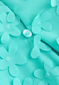 Türkise kangas, millel on suured, kihilised lille appliikatsioonid; keskel on ümar, helesinine nööp; sile tekstuur.