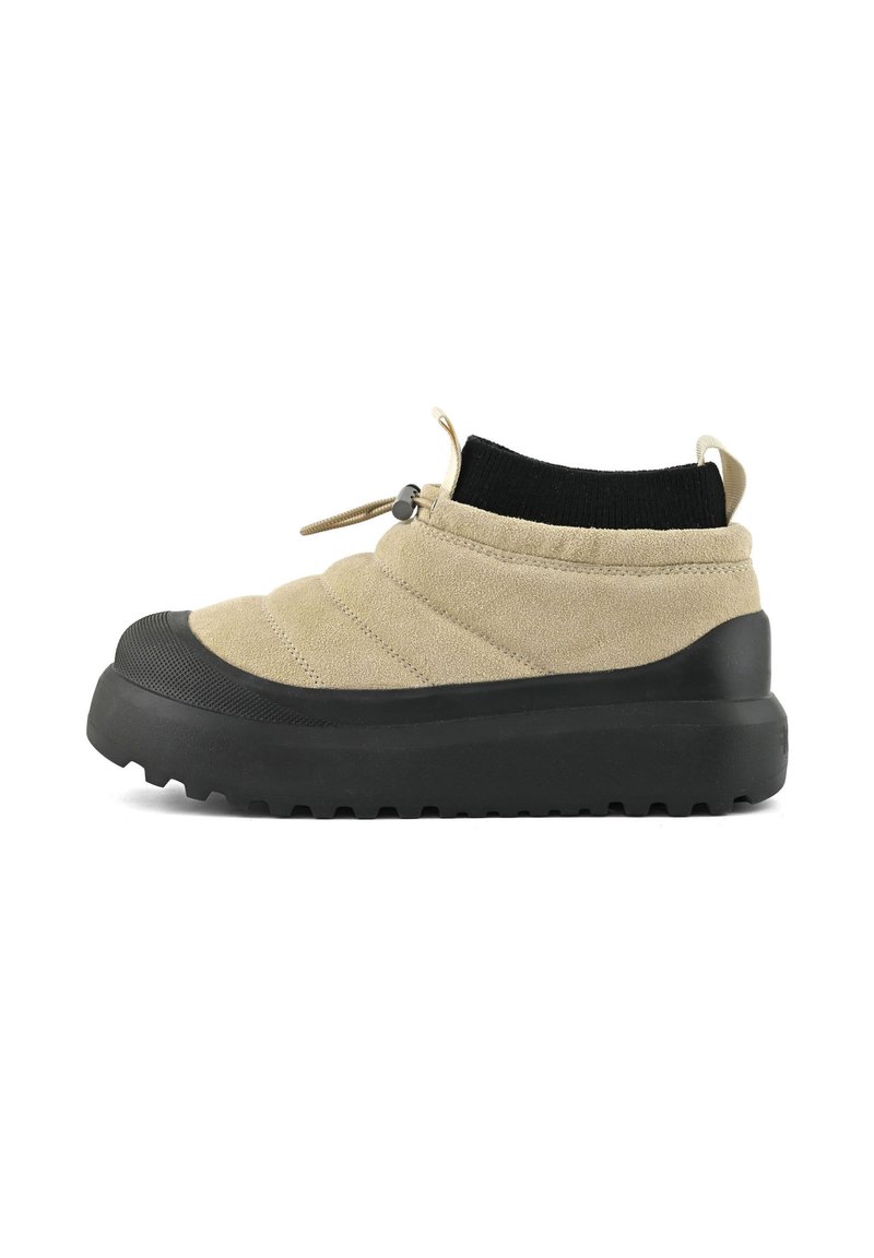 Beige slip-on tenisky s černou silnou gumovou podrážkou, žebrovaným černým nápletem na kotníku a nastavitelným stahováním na bílém pozadí.