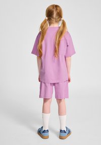 Lila Baumwoll-T-Shirt und Shorts-Set mit lockerer Passform, kombiniert mit weißen Socken und blauen Turnschuhen. Haare in zwei Zöpfen gebunden.