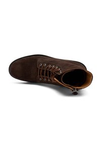 Bota de ante marrón con punta redondeada, cierre con cordones en la parte delantera y un forro interior en contraste. Presenta ojales plateados y una suela de goma negra.