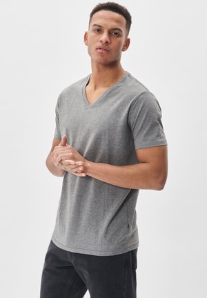 Camiseta de algodón gris con cuello en V y mangas cortas, que presenta un corte relajado y detalles en las costuras laterales, combinada con jeans oscuros.
