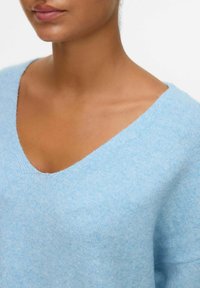 Pull en maille bleu clair avec un décolleté en V, texture douce et manches courtes, mettant en avant une coupe décontractée et de subtiles variations de teinte.