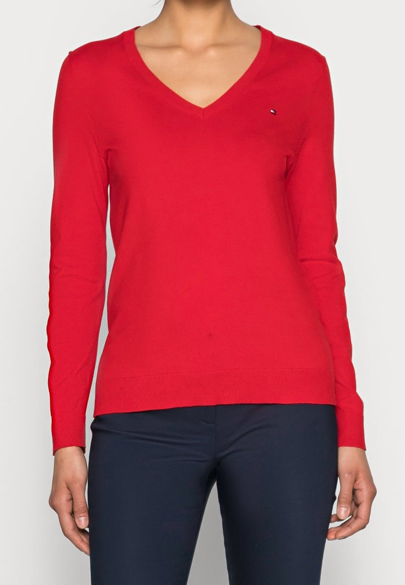 Femme portant un pull rouge ajusté à col en V et manches longues et un pantalon bleu marine foncé, visible du cou jusqu'aux hanches, debout devant un fond uni.