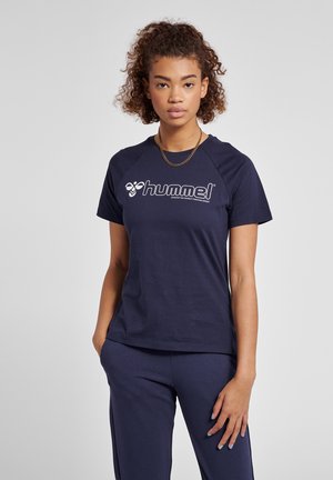 Junge Frau mit lockigem Haar, die ein navyblaues Hummel-T-Shirt und passende Hose trägt, steht mit einer Hand in der Tasche vor einem grauen Hintergrund.
