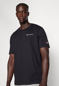 Sort bomulds T-shirt med korte ærmer, der har et hvidt broderet logo på brystet og et rødt og hvidt logo på ærmet.
