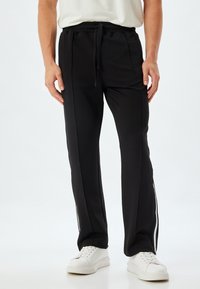 Pantalones deportivos negros con cintura elástica ajustable, bolsillos laterales y rayas blancas contrastantes a lo largo de las costuras exteriores. Tejido suave, corte ajustado.
