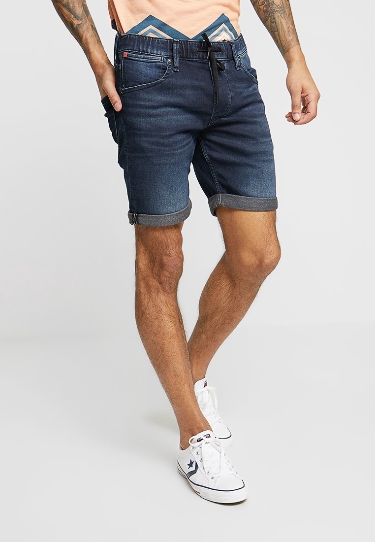Jeansshorts i mörkblått med upprullade avslut, har bälteshällor och sidofickor, matchade med vita sneakers. Slät tygtextur.