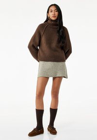 Kvindelig står iført en brun polo sweater, grå mini-skirt, mørke knæhøje sokker og brune loafers foran en ensfarvet hvid baggrund.