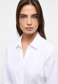 Camicia bianca con bottoni e colletto, realizzata in tessuto testurizzato e dal design aderente. Nessun motivo o accento visibile. Stile pulito e minimalista.