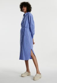 Himmelblaues, knöpfbares Kleid mit tailliertem Bindegürtel, langen Ärmeln und seitlichen Schlitzen. Tragen mit beigen Clogs. Strukturierter Stoff mit lockerer Passform.