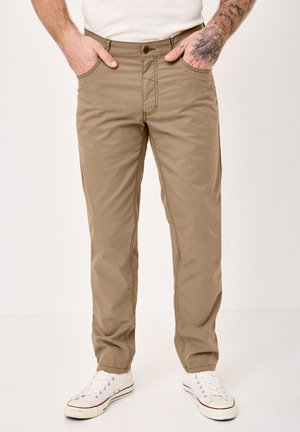 Man wearing beige straight-fit pants with side and front pockets, white sneakers, and tattooed forearms, hands in pockets.