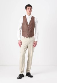 Rudas houndstooth liemenė su šešiais mygtukais, balta marškiniai, grietinėlės spalvos kelnės ir juodi batai. Medžiagos atrodo tekstūruotos ir pritaikytos.