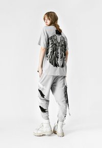 Camiseta gris de gran tamaño con diseño de alas de lentejuelas negras en la parte posterior, combinada con pantalones grises que presentan patrones de plumas en blanco y negro, y botas blancas.