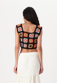 Háčkovaný crop top v čiernej farbe so živými kvetinovými štvorcami. Štvorcový výstrih, široké ramienka a textúrovaná látka. Nosený s bielou sukňou.